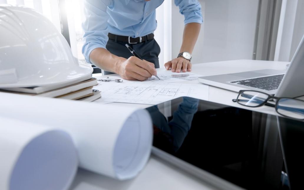 A man in business casual clothing works on construction plans. A hard hat, laptop and several rolls of paper sit around the plans on a table