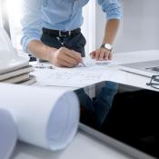 A man in business casual clothing works on construction plans. A hard hat, laptop and several rolls of paper sit around the plans on a table