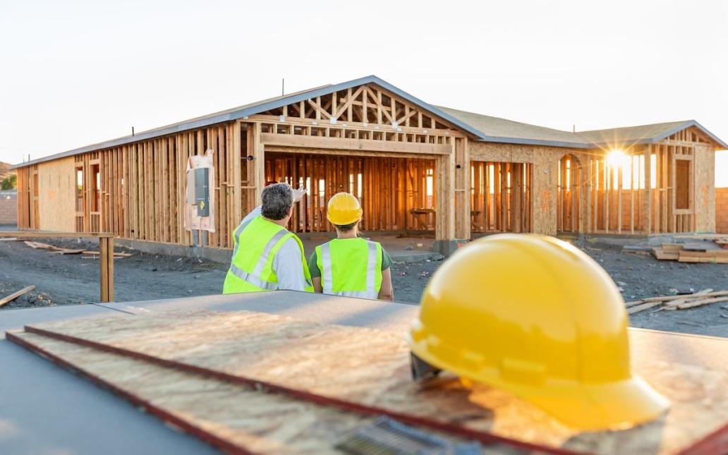 Two home builders stand facing under-construction home. A hard hat sits in the foreground. 
