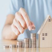 An individual sits at a table, stacking rows of coins in height ascending order. A wooden model home sits next to the coins, indicating home price growth. 