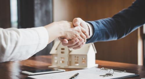 Two individuals sit at a desk and shake hands. On the desk, there is a set of keys, a model home and documentation, indicating a home sale. 