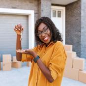 A woman stands in front of a house while smiling and holding up house keys. There are boxes in the driveway, indicating that she is moving in.