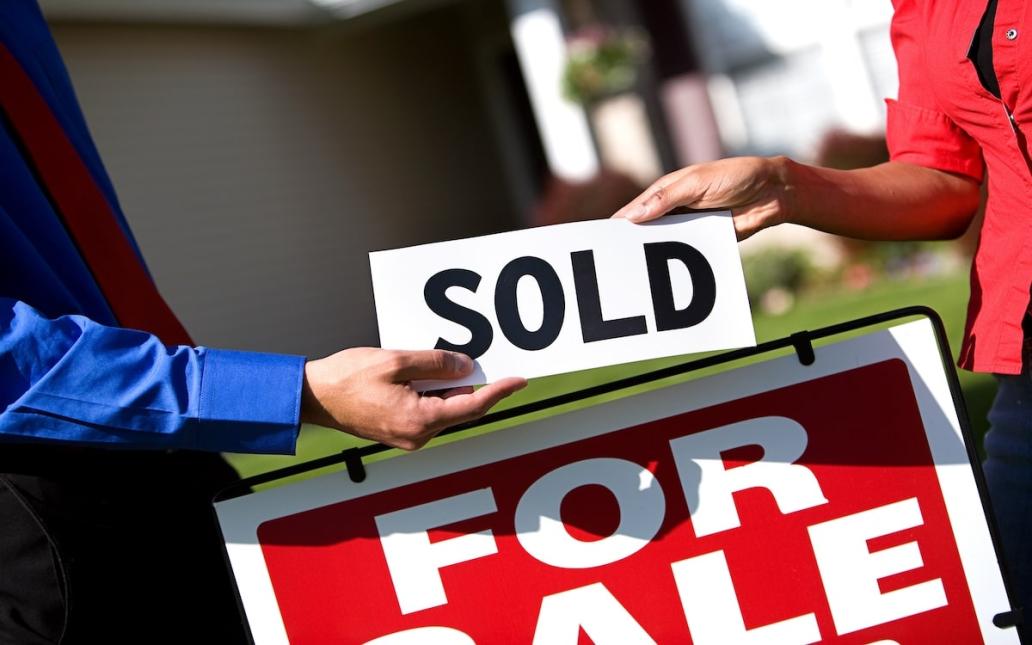 One person hands another person a sign reading "sold." A "For Sale" sign is in the background.