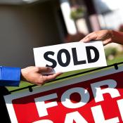 One person hands another person a sign reading "sold." A "For Sale" sign is in the background.